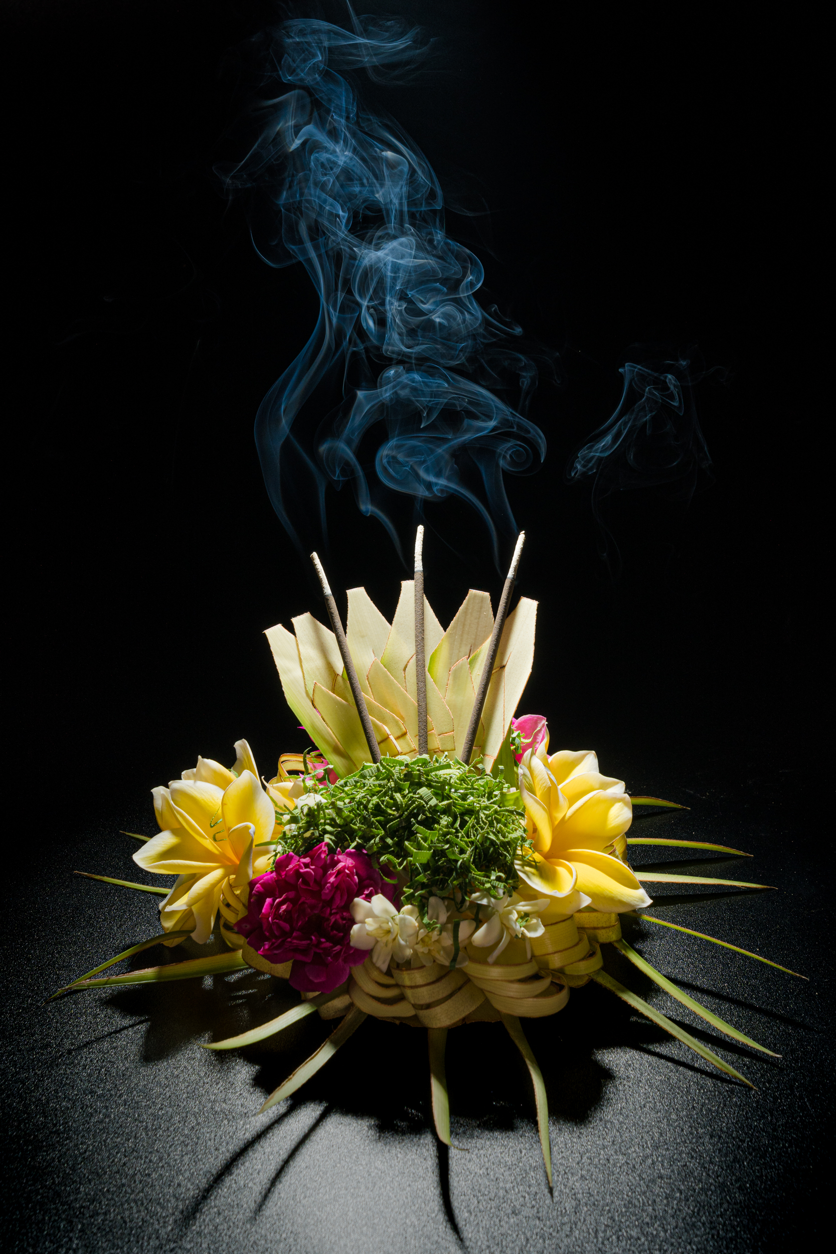Flowers and woven palm leaves in a canang sari offering, with incense smoke against a dark background